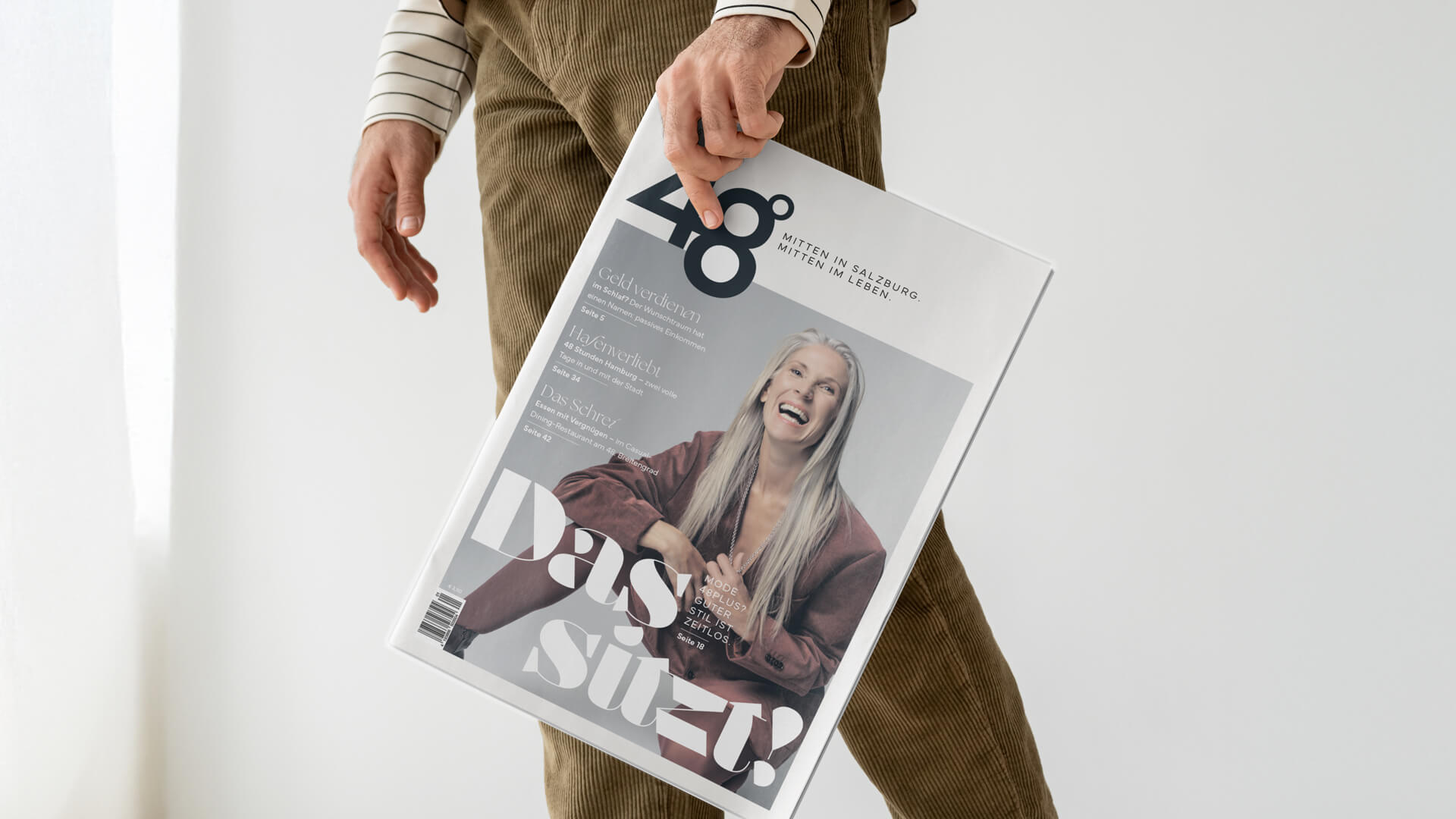 Eine Person hält eine Zeitschrift mit der Nummer 48 und einem Titelbild, das eine Frau mit einem Schal zeigt. Der Hintergrund ist weiß, und die Person trägt eine braune Jacke. Die Zeitschrift scheint sich mit Mode oder Lifestyle zu beschäftigen.