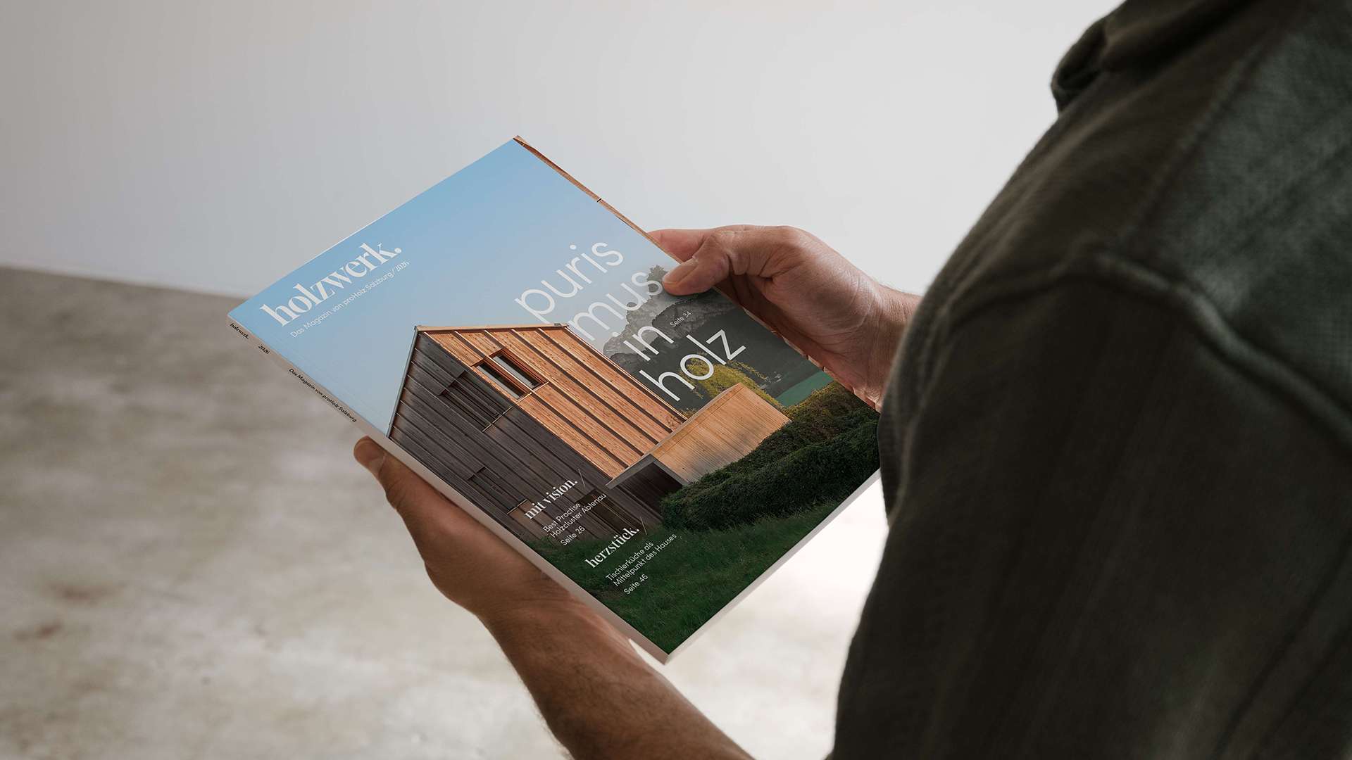 Eine Person hält ein Magazin oder eine Broschüre mit einem Cover, das ein Haus und den Text "Das Haus im Wald" zeigt. Der Hintergrund ist ein Raum mit grauer Wand und Betonboden.