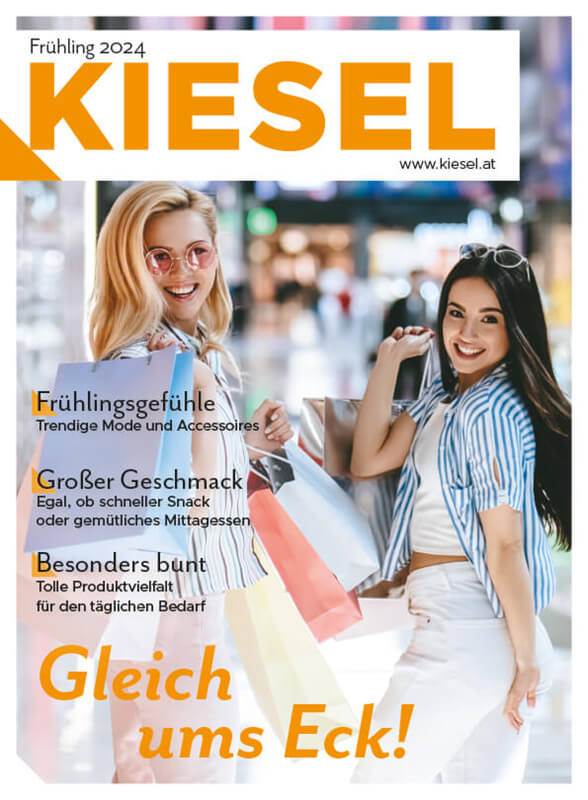 Zwei lächelnde Frauen halten jeweils eine Packung Kiesel Fruchtgummis in den Händen. Die Verpackungen zeigen bunte Fruchtgummis. Im Hintergrund sind weitere Produkte und ein Regal mit Süßigkeiten zu sehen.